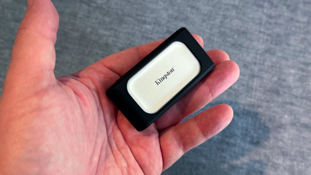 A hand holding a small rectangular device with a black border and a white center featuring the word Kingston.